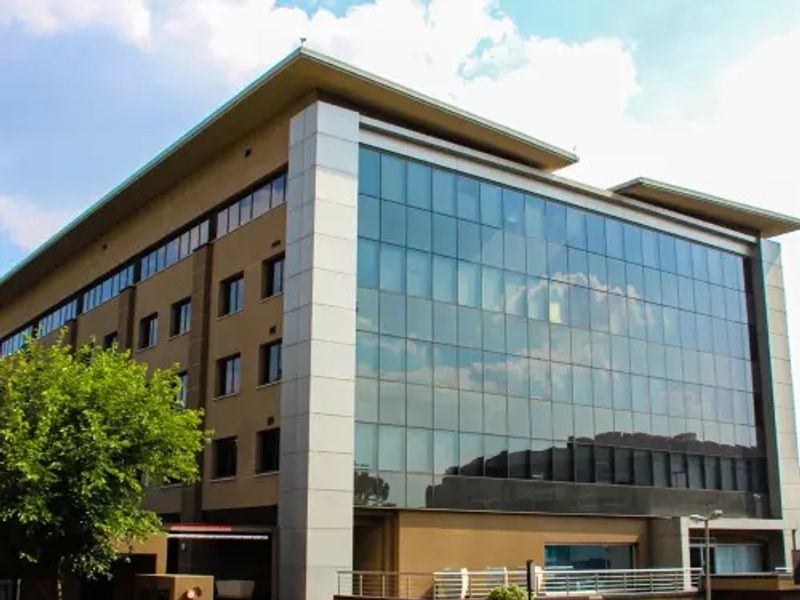 Modern commercial office building featuring a prominent glass curtain wall facade, metal cladding, and a paved driveway with secure access.