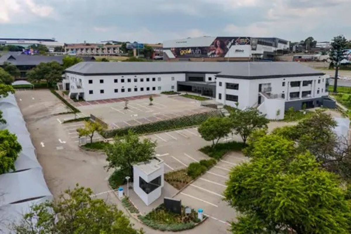 Aerial view of a commercial property featuring a multi-story building, extensive paved parking area, security guard house, and landscaped grounds.