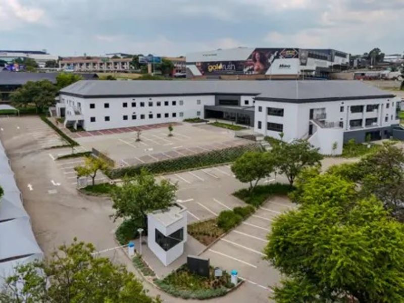 Aerial view of a commercial property featuring a multi-story building, extensive paved parking area, security guard house, and landscaped grounds.