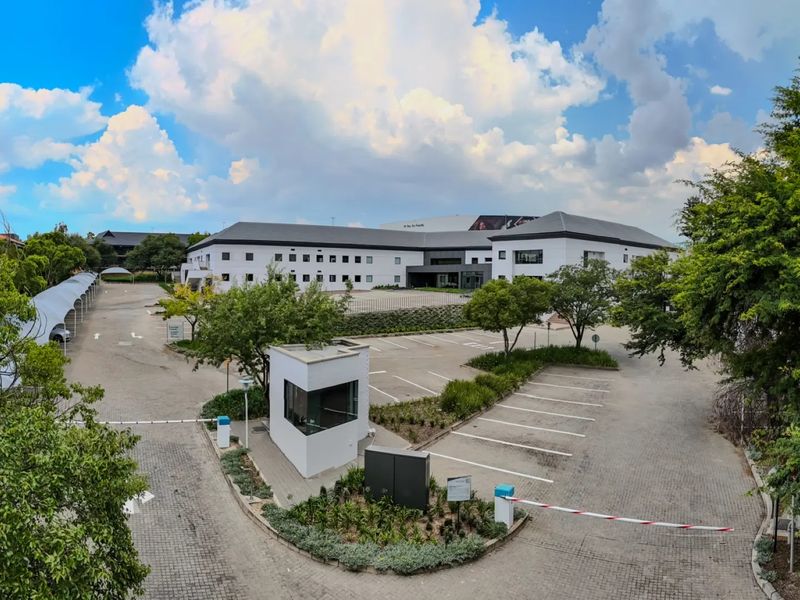 Wide-angle view of a commercial property featuring a secure gated entrance, paved parking lot, covered parking bays, and a multi-story office building.
