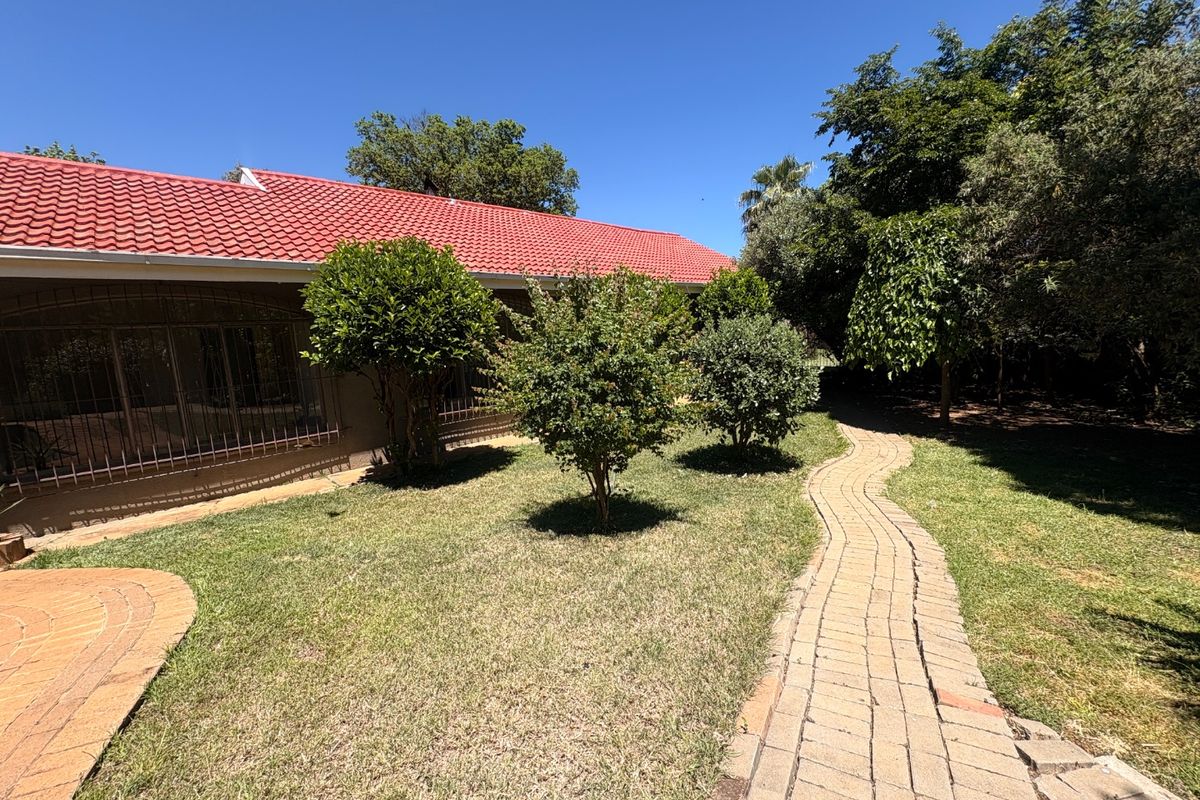 A rear garden view featuring a paved walkway, established trees, and a house exterior with a red tiled roof and security bars.