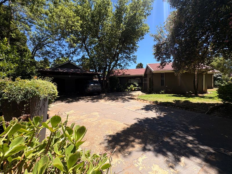 Front view of the property featuring a paved driveway, carport, and established garden with mature trees under a tiled roof.