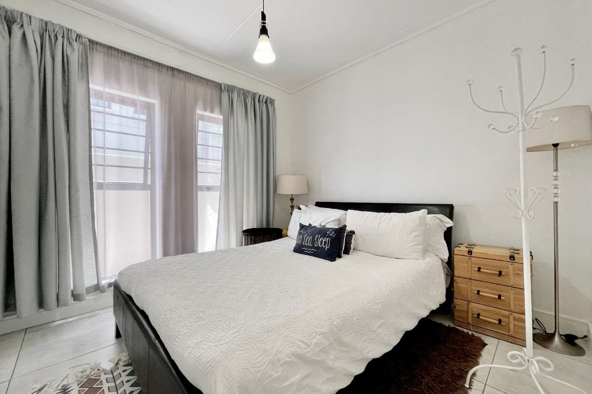 A well-lit bedroom featuring tiled flooring, neutral walls, and large windows fitted with security bars and curtains.