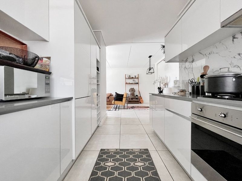 Modern galley-style kitchen featuring sleek white cabinetry, stone countertops, integrated oven, and a marble-look splashback, leading into an open-plan living area.