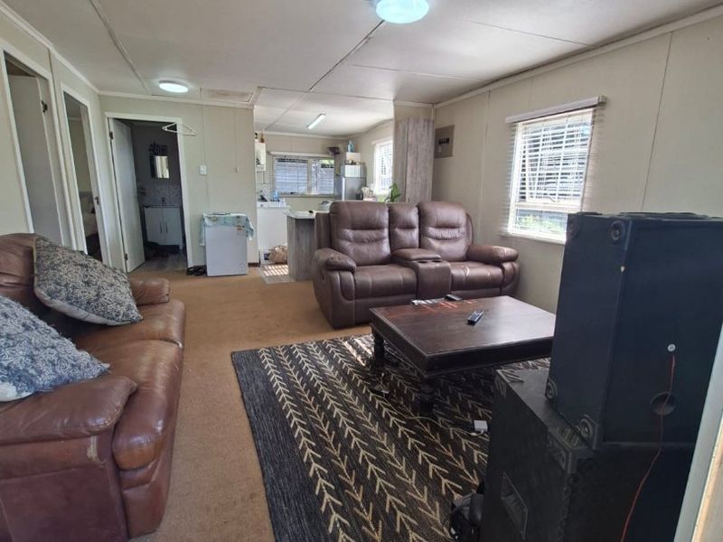 Open plan living area featuring dated wall paneling, carpeted flooring, and ceiling tiles, with an adjacent kitchen area visible in the background.