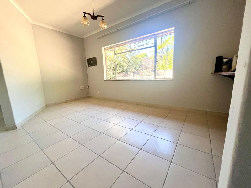 A bright, empty living room featuring neutral tiled flooring, a large window with security bars, and a central ceiling light fixture.