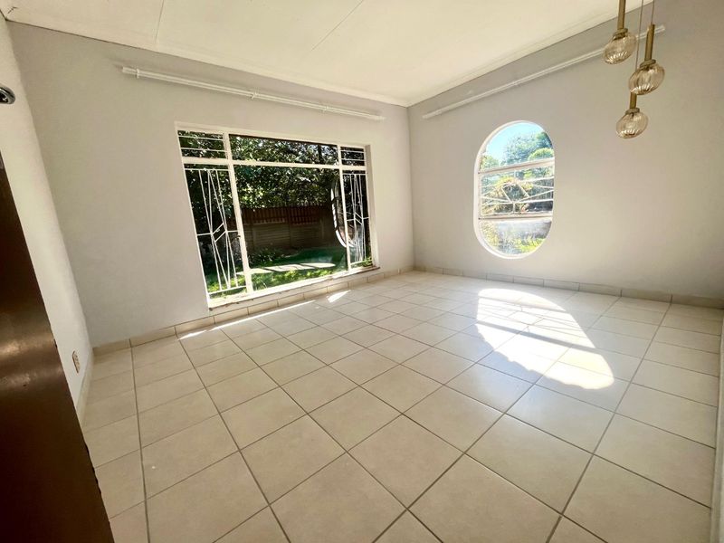 A bright, empty living room featuring ceramic floor tiles, a large rectangular window, and a distinctive oval window with security bars.