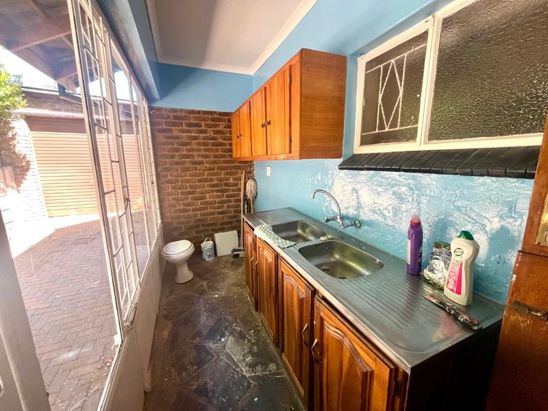 A functional scullery area featuring wooden cabinetry, a double stainless steel sink, and exposed brickwork, providing practical utility space with direct outdoor access.