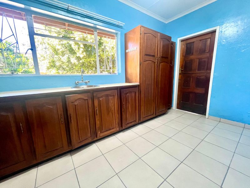 A functional kitchen area featuring wooden cabinetry, a stainless steel sink, tiled flooring, and a secure window with burglar bars.