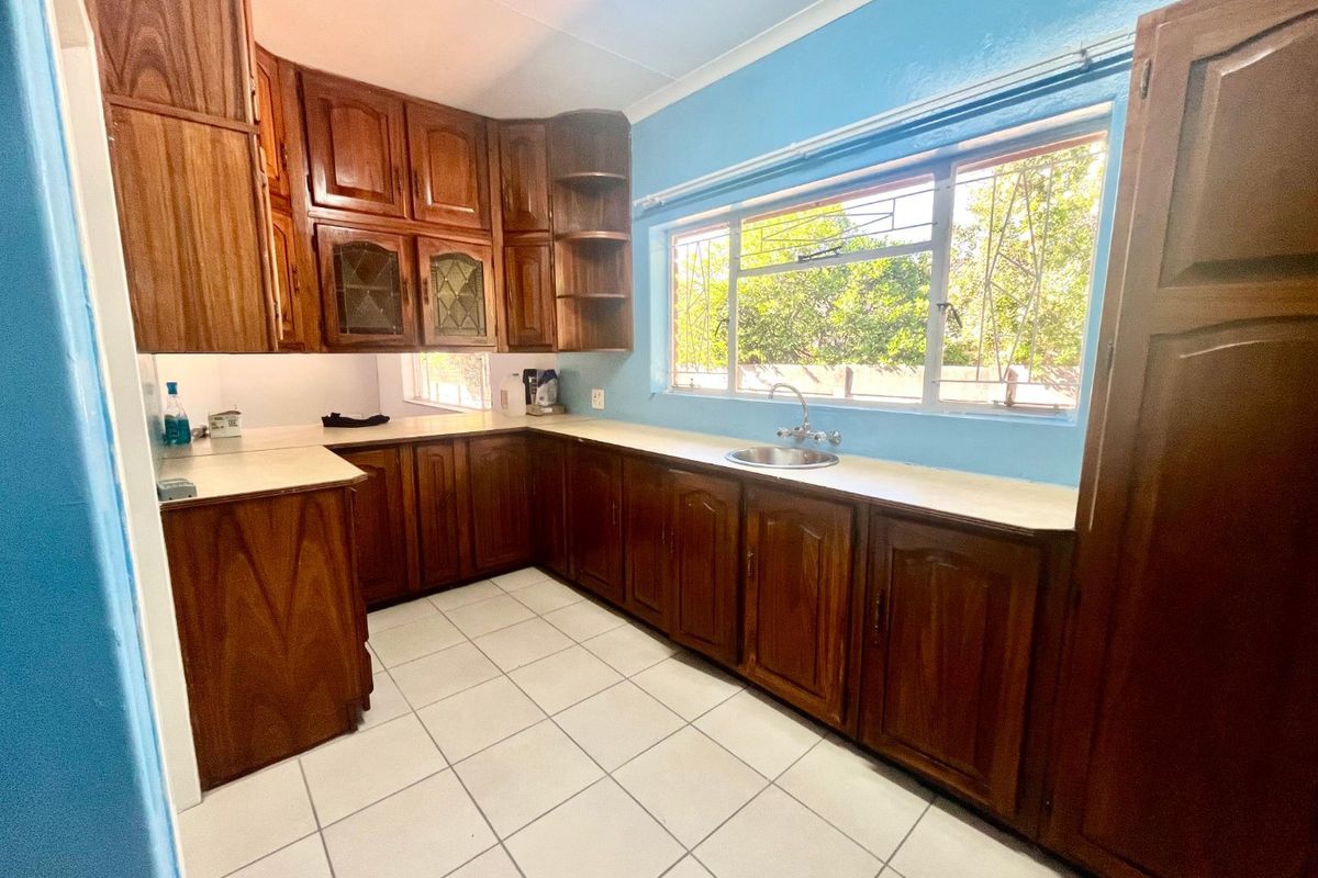 A functional kitchen featuring traditional wooden cabinetry, laminate countertops, a stainless steel sink, and tiled flooring with natural light from a large window.