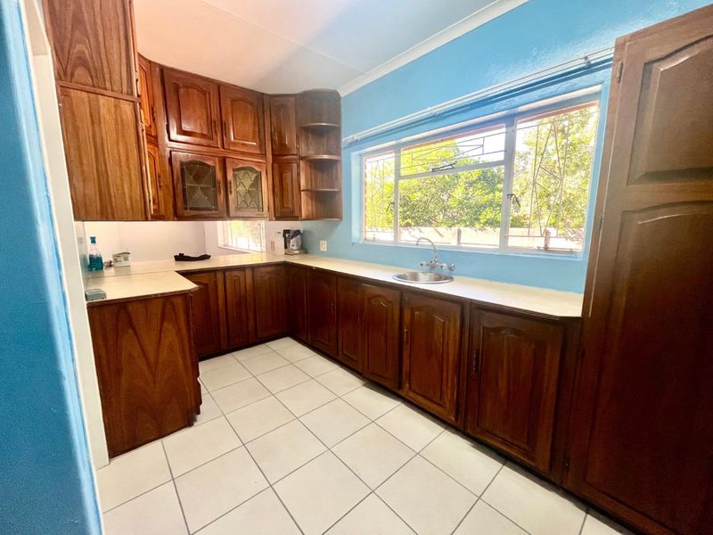 A functional kitchen featuring traditional wooden cabinetry, laminate countertops, a stainless steel sink, and tiled flooring with natural light from a large window.