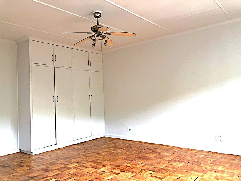 A bright, empty bedroom featuring classic parquet flooring, ample built-in cupboard storage, and a ceiling fan.