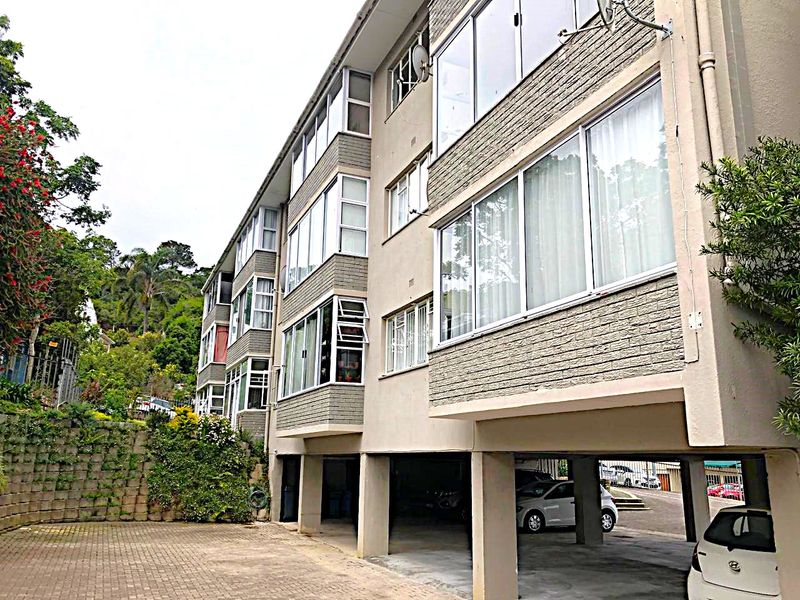 Exterior view of a multi-story apartment building featuring brick cladding, large sliding windows, and ground-level undercover parking with a paved driveway.