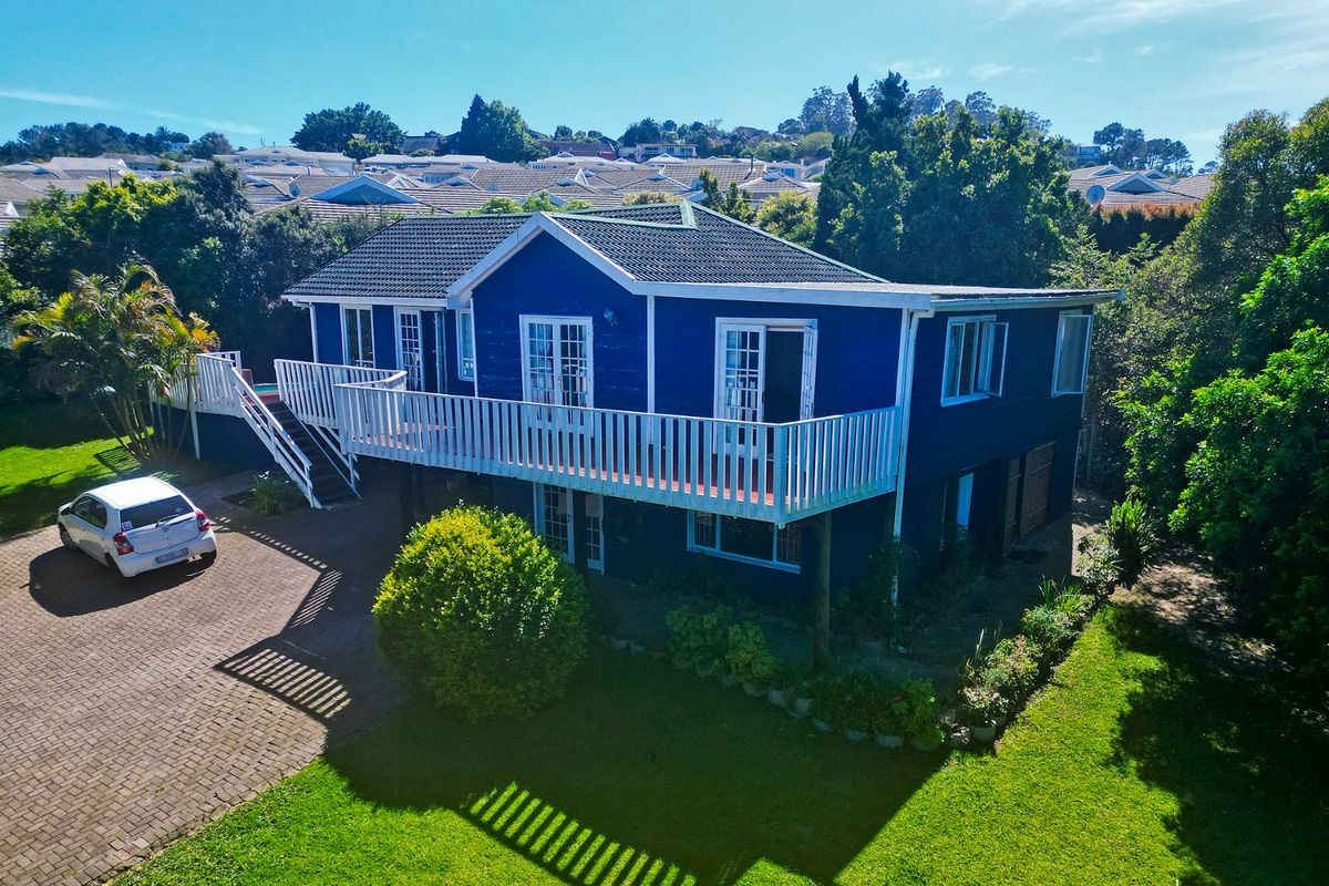Aerial view of a blue-painted detached house featuring an elevated deck, paved driveway, and surrounding garden in a residential neighborhood.
