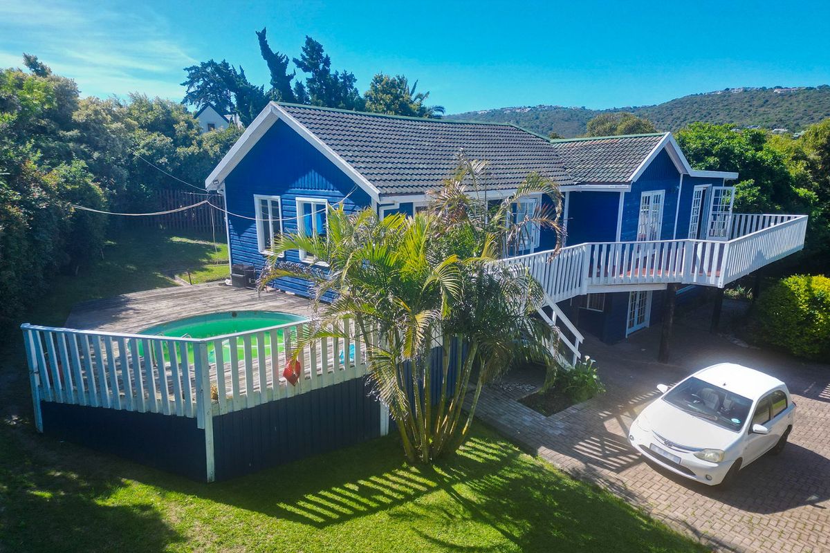 Rear view of a blue timber-clad house featuring a raised wooden deck, swimming pool, and paved driveway area.