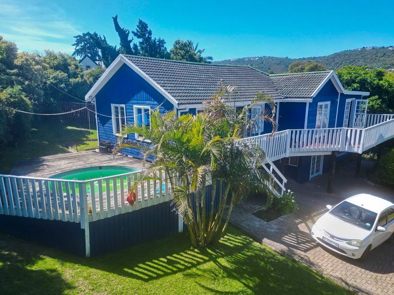 Rear view of a blue timber-clad house featuring a raised wooden deck, swimming pool, and paved driveway area.