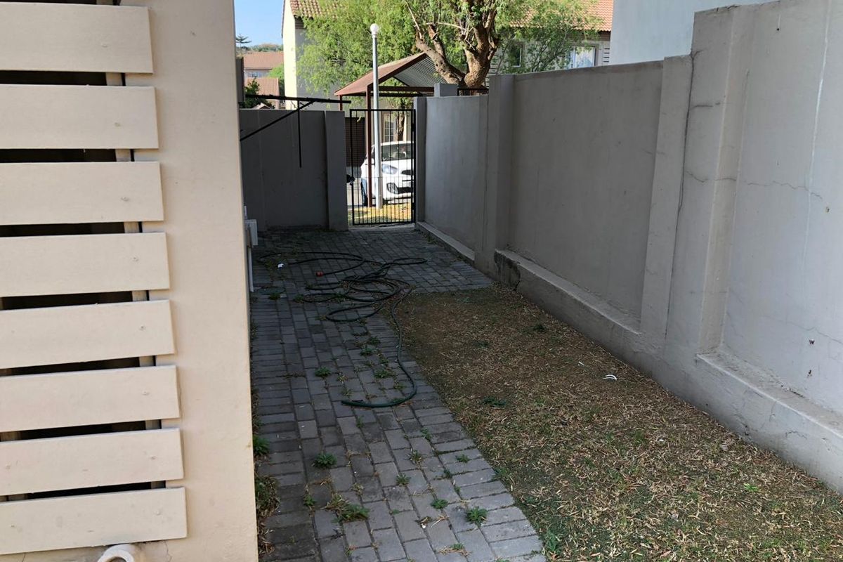 A narrow rear exterior area featuring a paved walkway, boundary wall, and metal security gate, requiring maintenance and landscaping improvements.