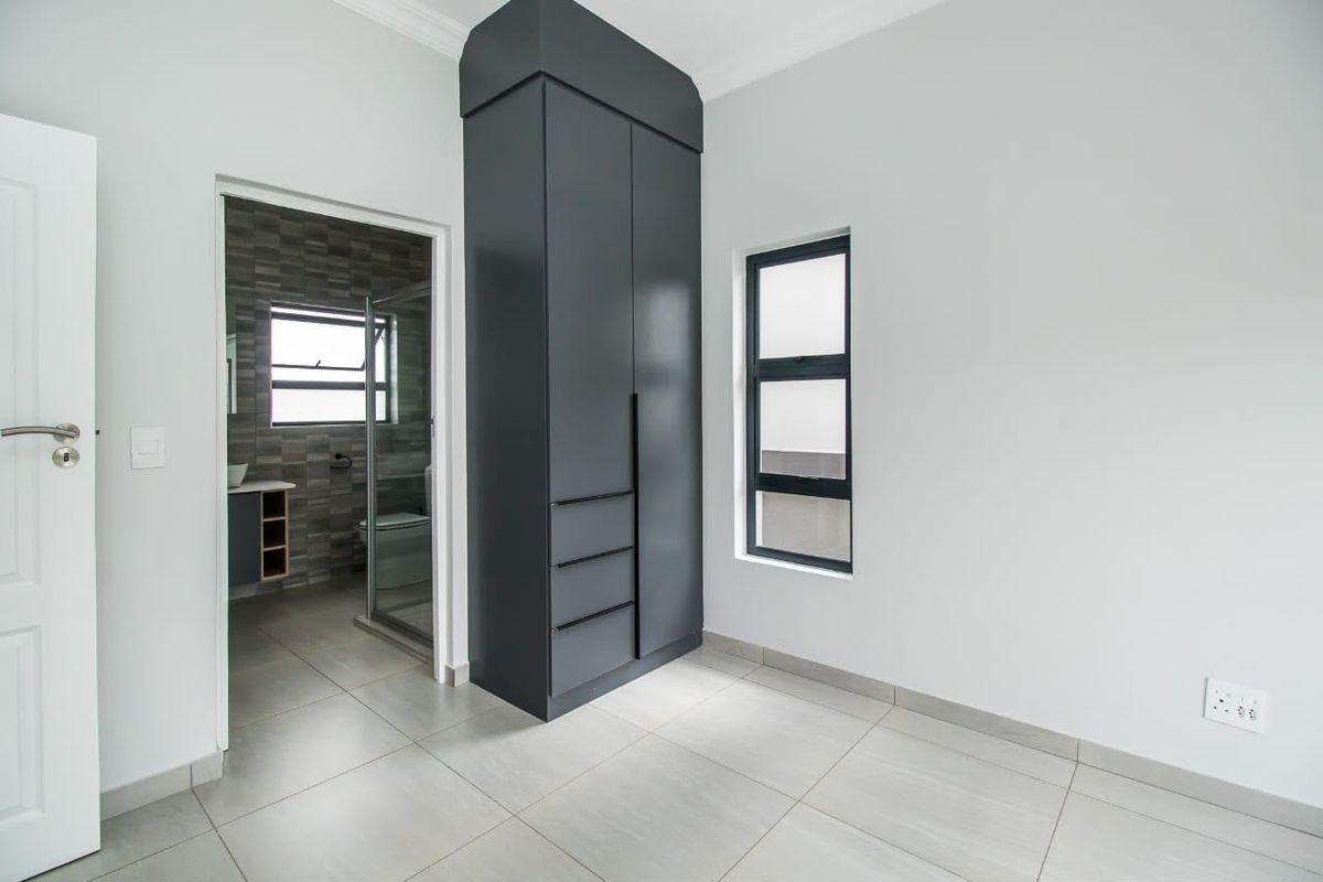 A clean, modern bedroom featuring neutral tiled flooring, a dark grey built-in cupboard, and an adjoining en-suite bathroom.