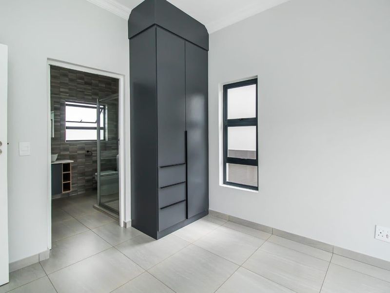 A clean, modern bedroom featuring neutral tiled flooring, a dark grey built-in cupboard, and an adjoining en-suite bathroom.