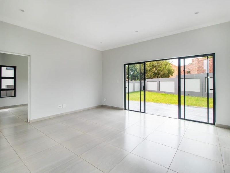 A bright, modern, and empty living room featuring large sliding glass doors that open onto a paved patio and garden area.