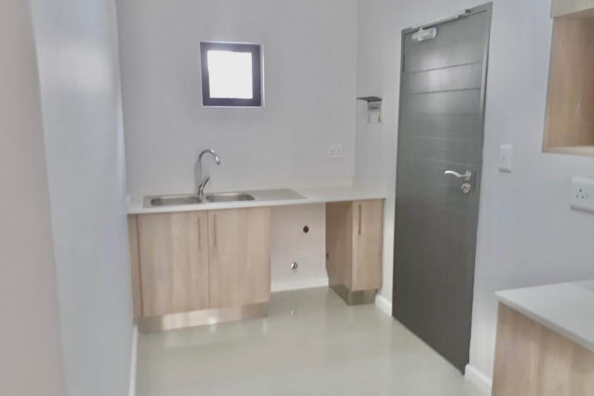 A clean, modern kitchen area featuring light wood-finish cabinetry, a stainless steel sink, and neutral tiled flooring in a bright, minimalist space.