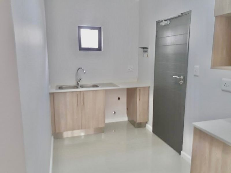 A clean, modern kitchen area featuring light wood-finish cabinetry, a stainless steel sink, and neutral tiled flooring in a bright, minimalist space.