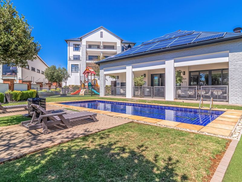 A well-maintained outdoor pool area featuring a tiled swimming pool, covered patio space, solar panels on the roof, and landscaped garden surroundings.