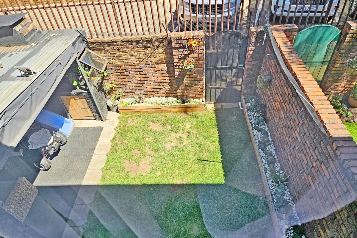 A private rear courtyard featuring a brick boundary wall, metal security gate, raised garden beds, and a small lawn area with a covered section.