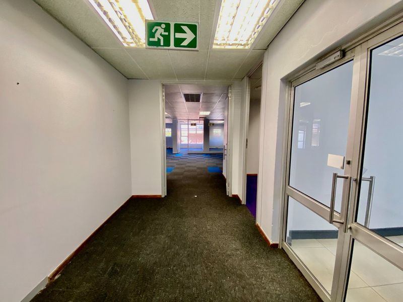 A commercial hallway featuring carpeted flooring, suspended ceilings with integrated lighting, and an aluminium-framed glass door leading to an open-plan office space.