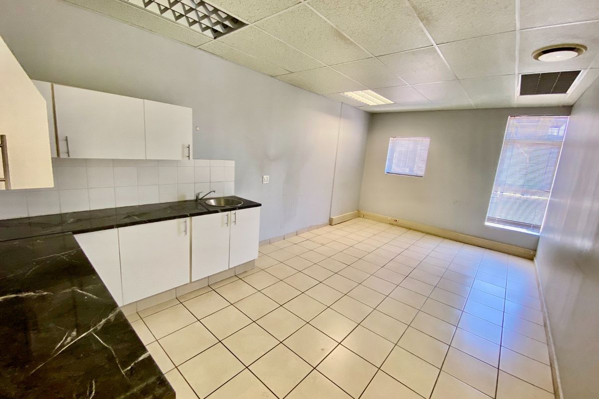 A functional commercial kitchenette featuring white cabinetry, dark laminate countertops, tiled flooring, and drop ceiling lighting in an open, vacant office space.