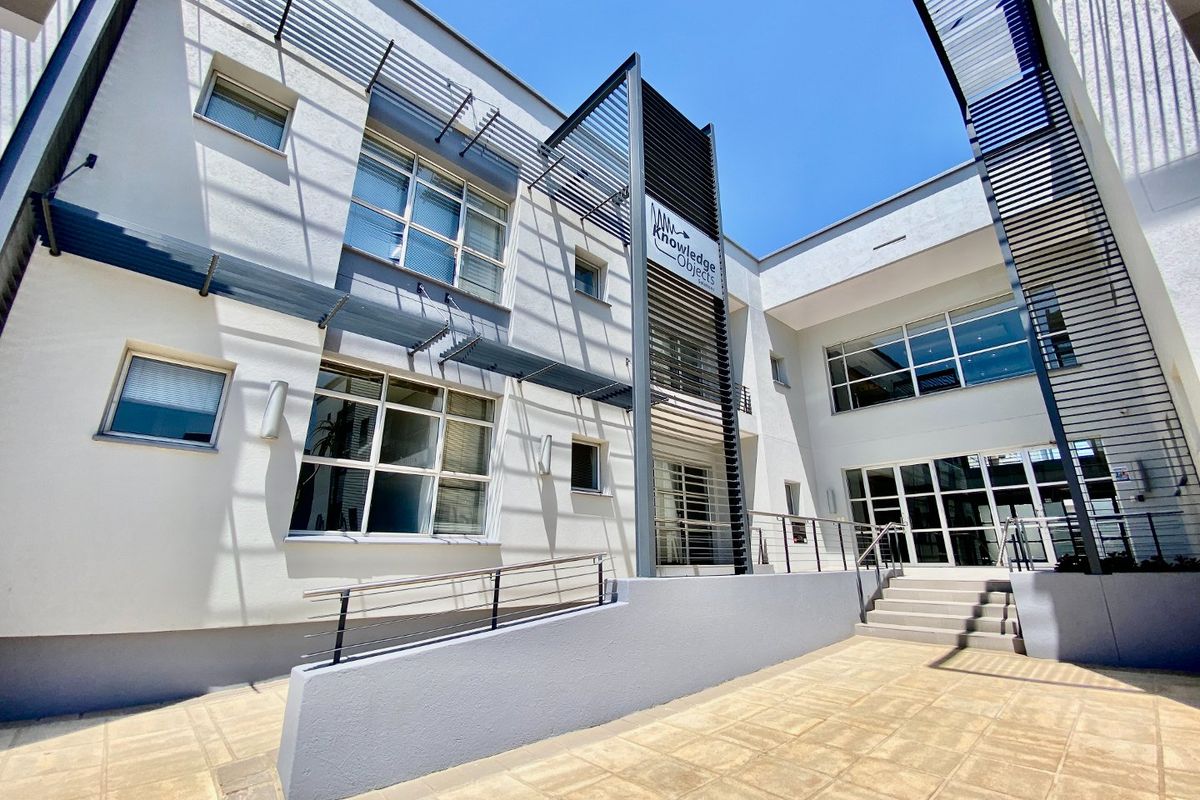 A modern commercial courtyard featuring paved flooring, an accessibility ramp with metal handrails, and architectural metal sunshades over large glass windows.