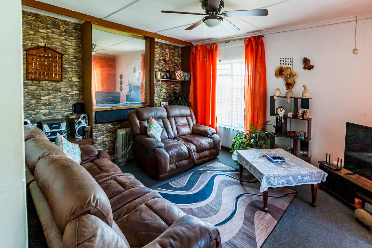 A living room featuring a stone accent wall, ceiling fan, and large window with security bars, offering a dated aesthetic ready for modernization.