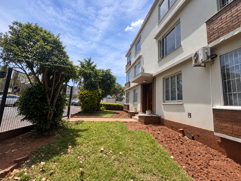 Front exterior view of an apartment building featuring a brick and plaster facade, security window bars, and a landscaped garden area.