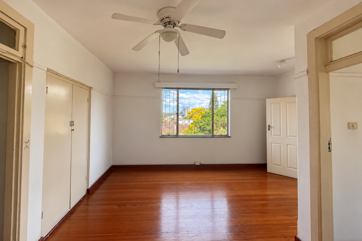 A bright, empty bedroom featuring polished timber flooring, built-in cupboards, a ceiling fan, and a window with security bars overlooking the neighborhood.