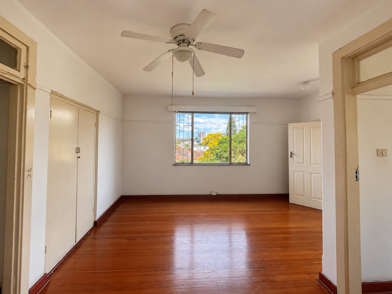 A bright, empty bedroom featuring polished timber flooring, built-in cupboards, a ceiling fan, and a window with security bars overlooking the neighborhood.