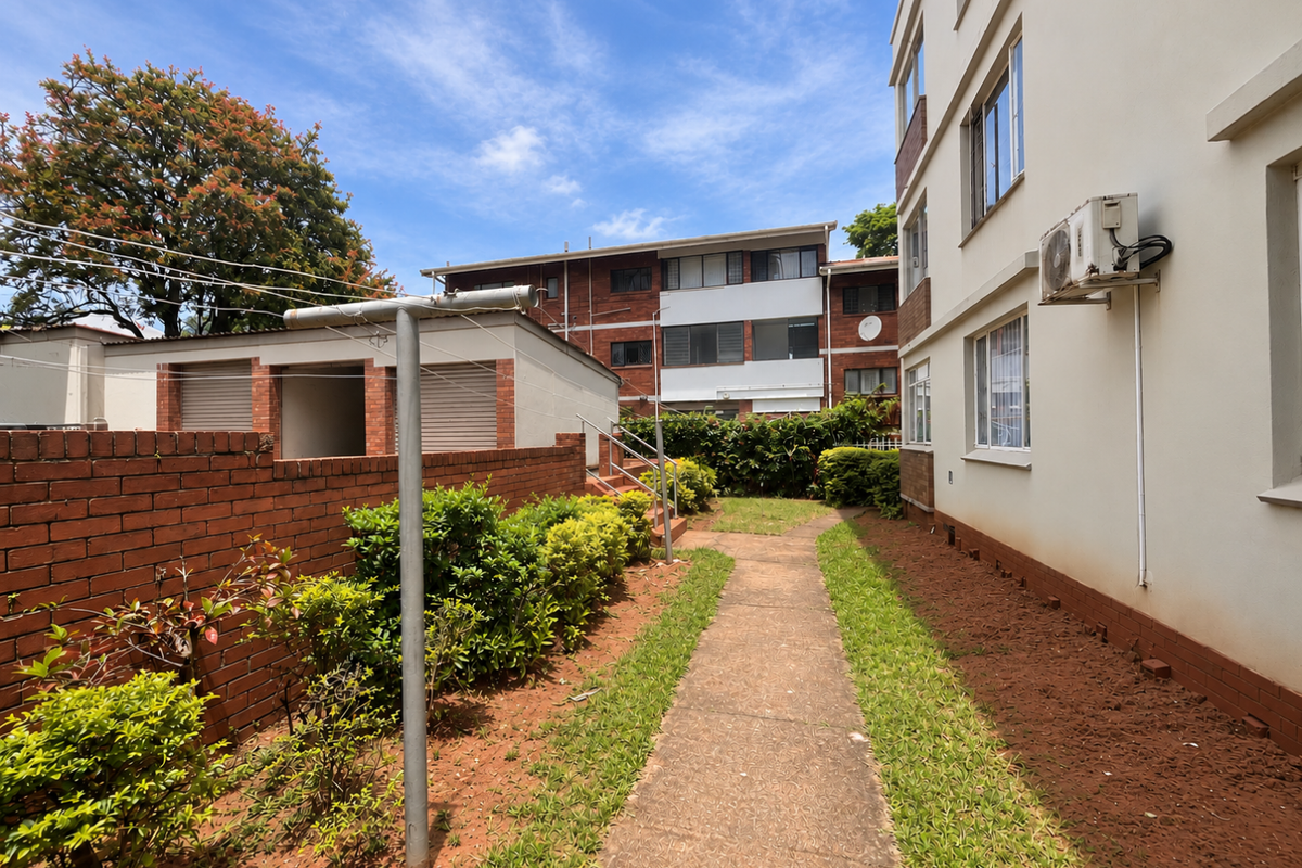 Rear exterior view of the apartment complex featuring a paved walkway, brick boundary wall, and established garden beds.
