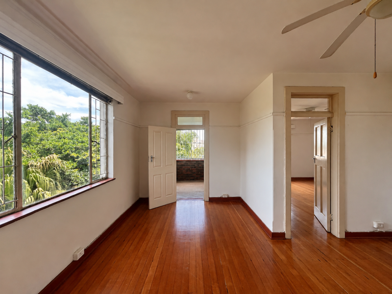 A bright, empty bedroom featuring polished hardwood floors, a ceiling fan, and large windows providing natural light and garden views.