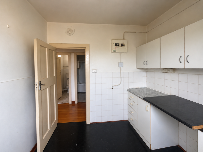 A compact, empty kitchen area featuring basic built-in cabinetry, tiled backsplash, and direct access to the bathroom and hallway.