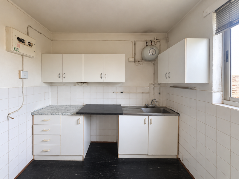 A functional, empty kitchen featuring white cabinetry, tiled splashbacks, a stainless steel sink, and exposed utility infrastructure, ready for modernization.