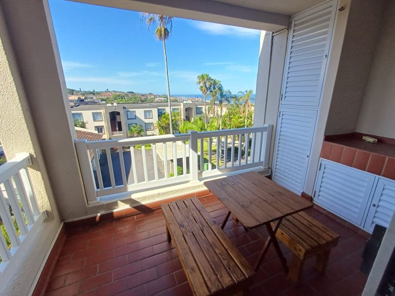 Covered balcony featuring a built-in braai, tiled flooring, and a white railing, offering pleasant views of the surrounding complex and distant ocean.