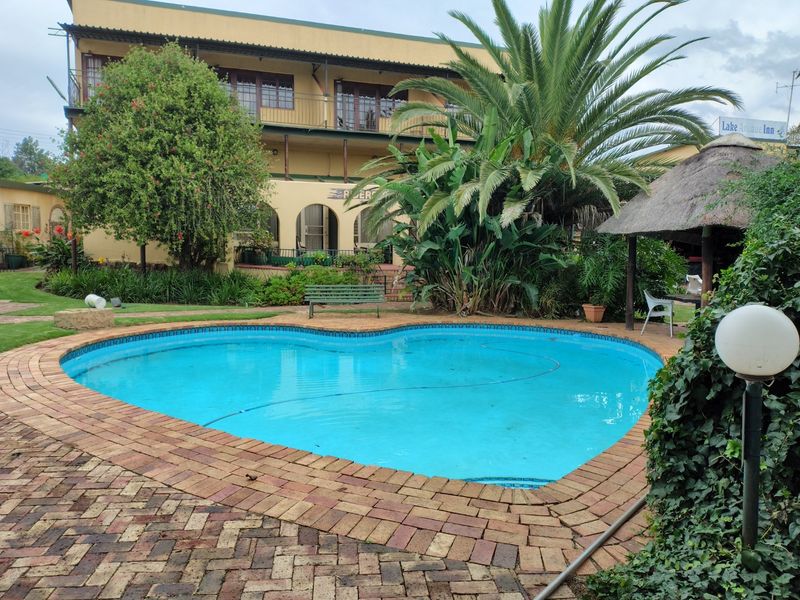 A heart-shaped swimming pool set in a landscaped garden with brick paving, a thatched lapa, and a multi-story building with a balcony in the background.