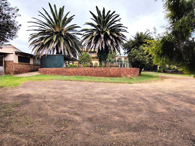 A wide view of the property frontage featuring a brick retaining wall, mature palm trees, a garden gazebo, and a large gravel driveway area.
