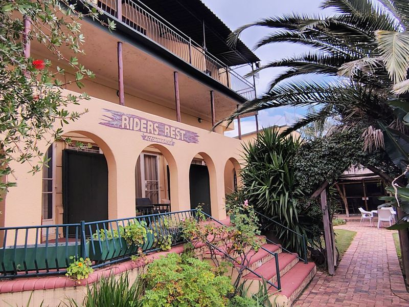 Exterior view of a multi-level building featuring an arched veranda, metal balcony railings, and a brick-paved pathway leading through a landscaped garden area.