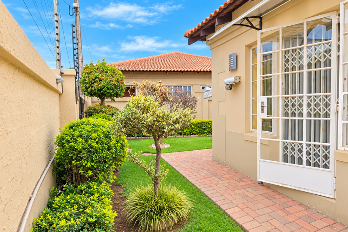 A well-maintained rear garden area featuring a brick paved walkway, manicured landscaping, and secure perimeter walling with an electric fence.