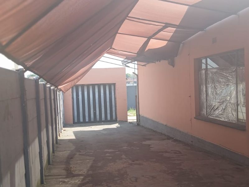 A covered outdoor side area featuring a concrete driveway, precast boundary wall, and a metal garage door, currently requiring maintenance to the canopy and finishes.