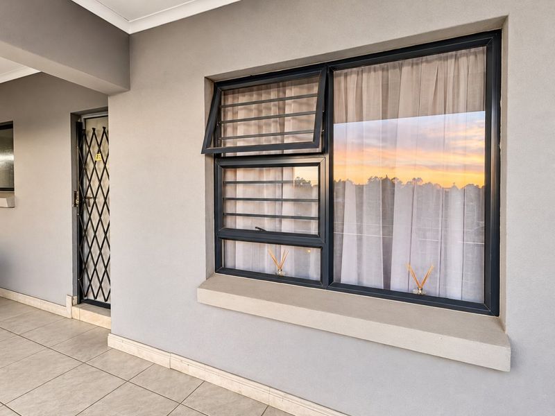 Exterior view of an apartment entrance featuring a security gate, tiled flooring, and aluminium window frames with security bars.