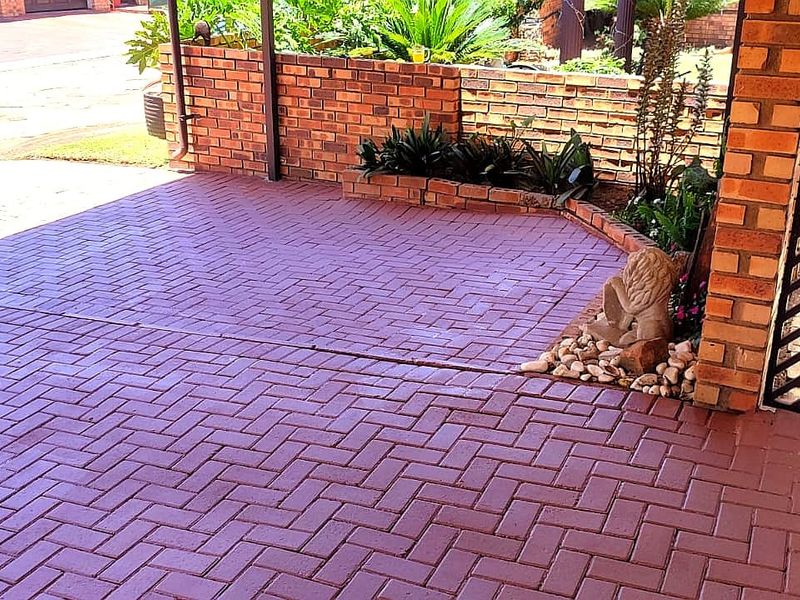 A covered patio area featuring herringbone brick paving, a low brick retaining wall with garden beds, and a secure metal entrance gate.