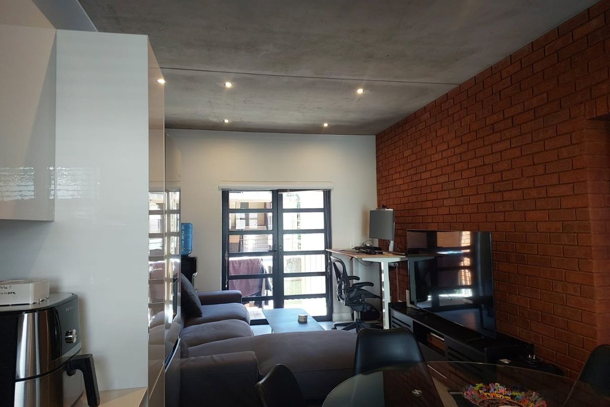 Open-plan living area featuring an exposed brick feature wall, concrete ceiling with recessed lighting, and modern high-gloss kitchen cabinetry leading to a patio.