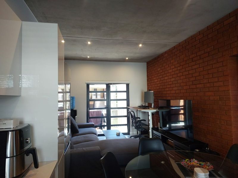 Open-plan living area featuring an exposed brick feature wall, concrete ceiling with recessed lighting, and modern high-gloss kitchen cabinetry leading to a patio.