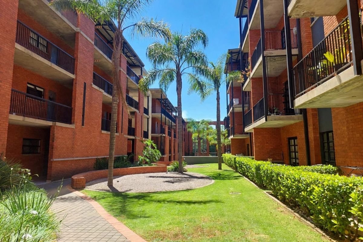A view of the apartment complex courtyard featuring exposed brick facades, private balconies with metal railings, and a well-maintained landscaped garden with paved walkways.